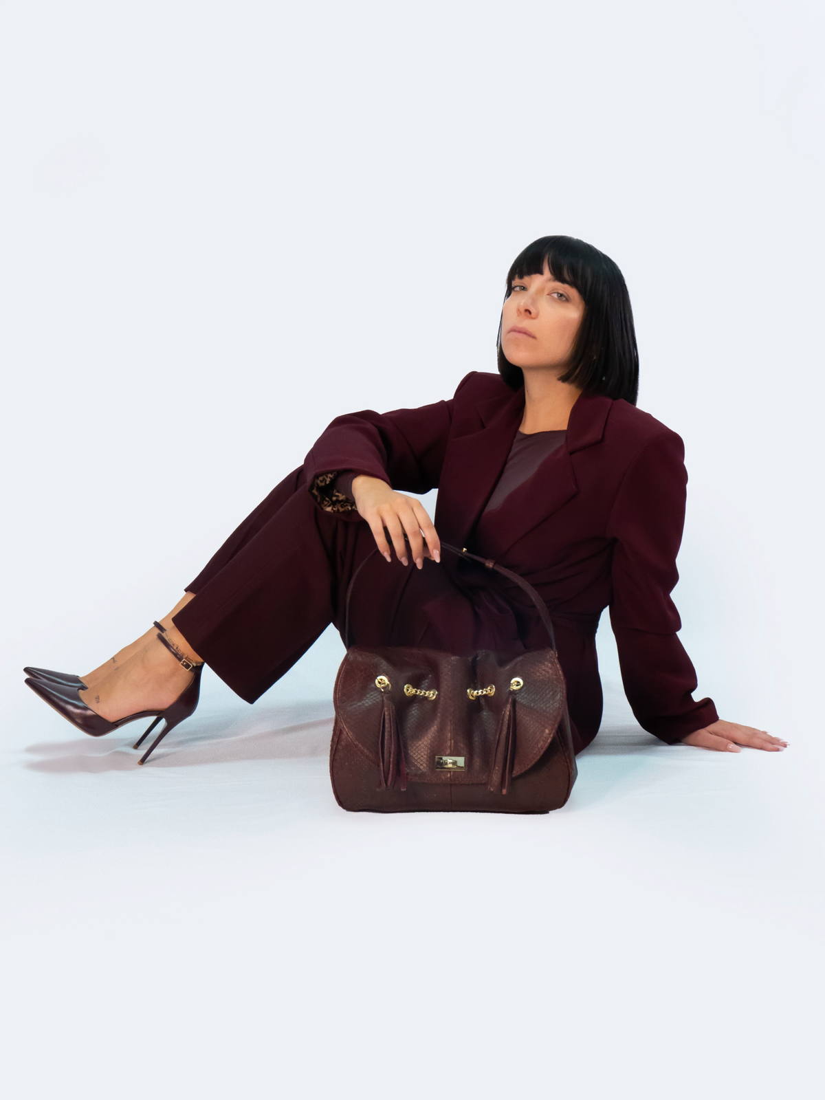 Modella seduta con borsa Amatì Onice in pitone bordeaux con dettagli dorati e nappe, abbinata a un completo elegante.